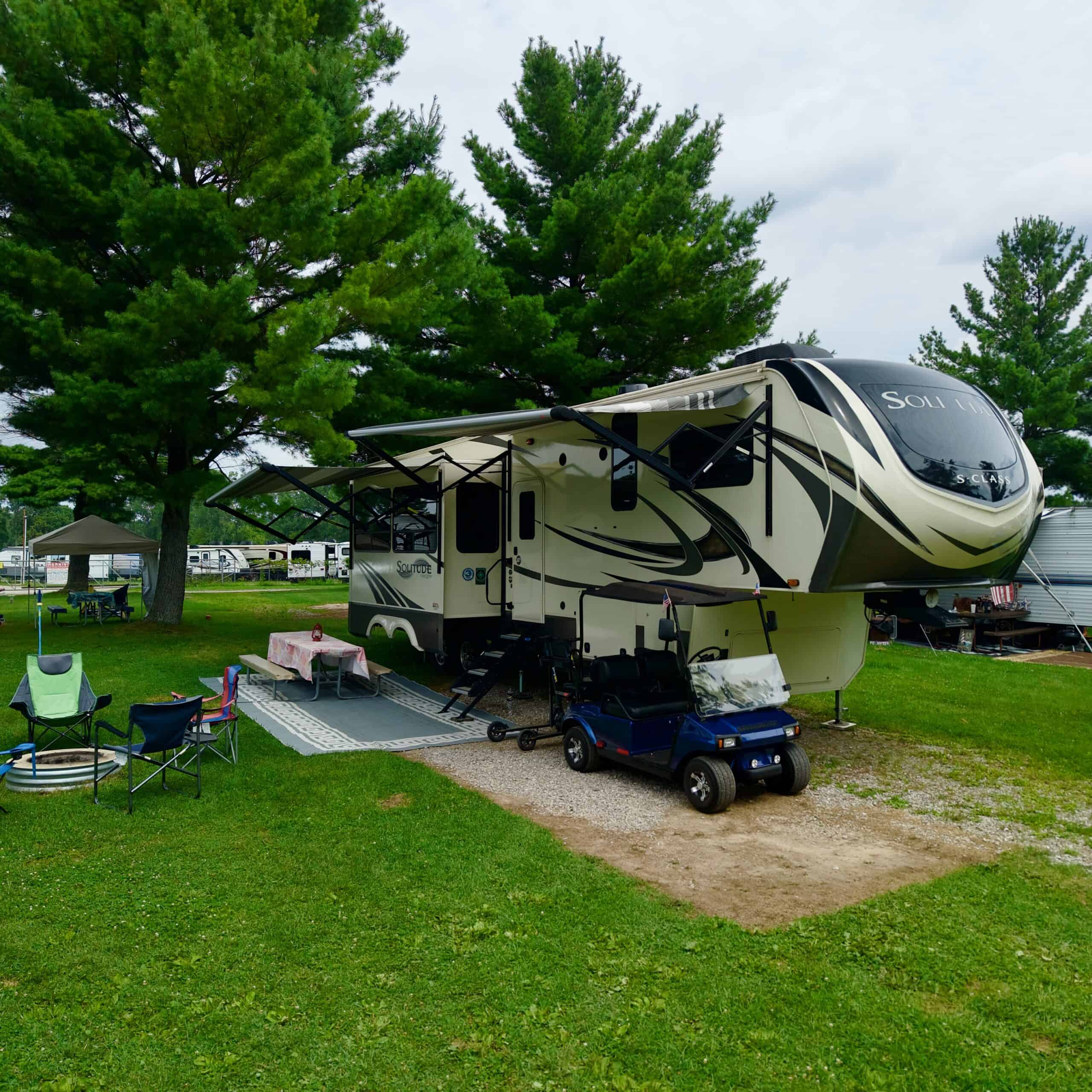 Spacious RV campsite under tall green trees with outdoor seating and a golf cart.